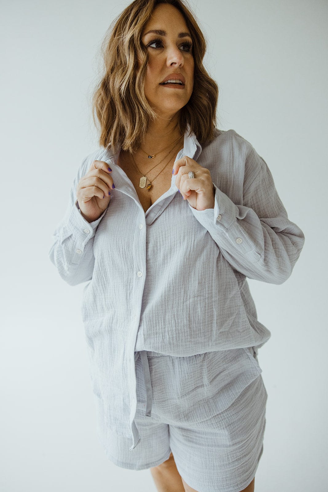 A woman with wavy hair wearing a Love Marlow SUPER SOFT TEXTURED BUTTON-UP BLOUSE in a light color, paired with shorts, is looking to her left against a plain white background.