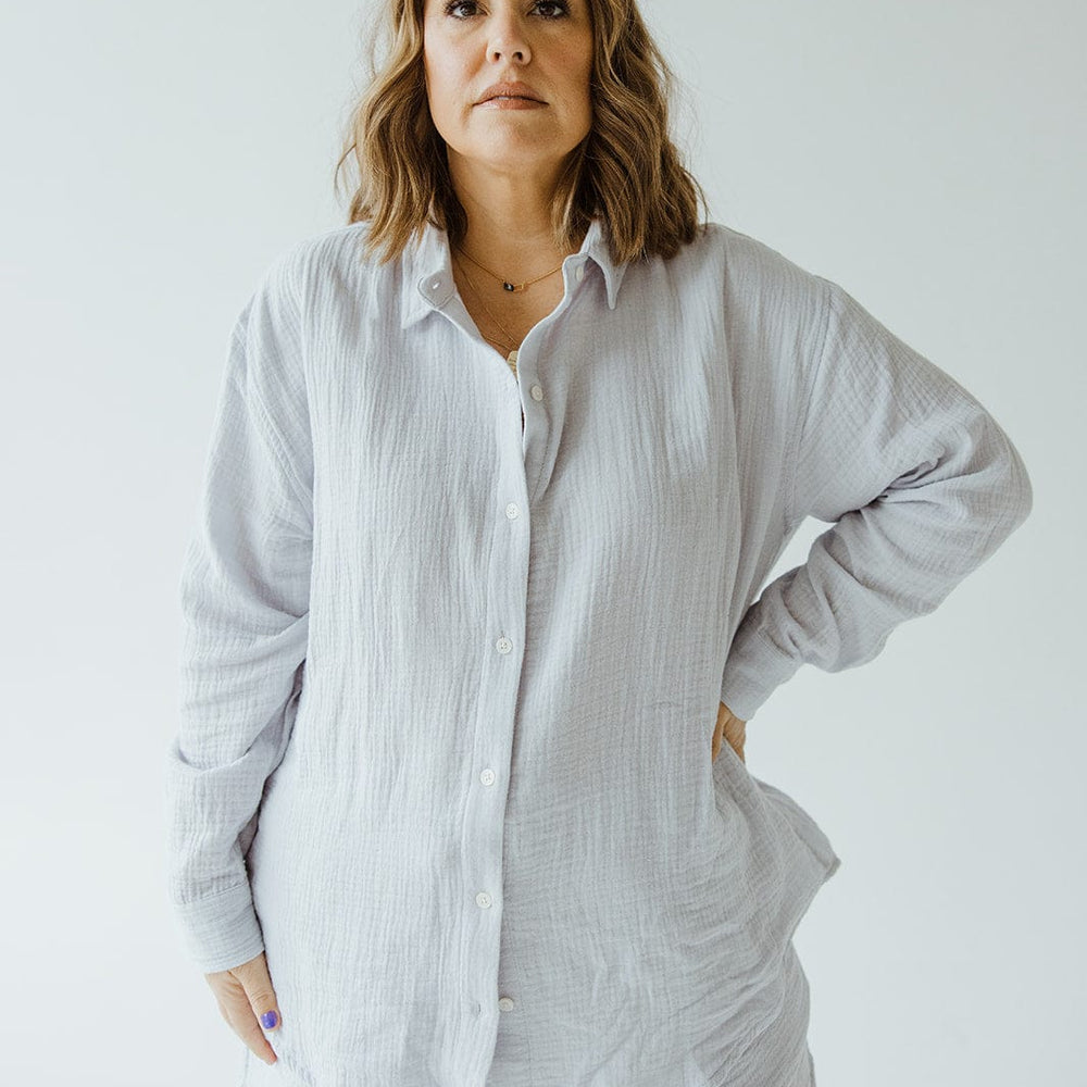 A person stands facing the camera with one hand on their hip, wearing a light-colored SUPER SOFT TEXTURED BUTTON-UP BLOUSE from Love Marlow and matching shorts.