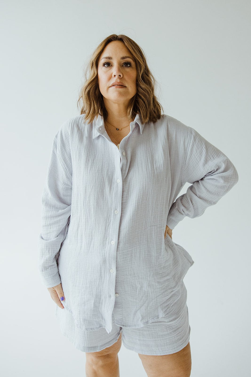 A person stands facing the camera with one hand on their hip, wearing a light-colored SUPER SOFT TEXTURED BUTTON-UP BLOUSE from Love Marlow and matching shorts.