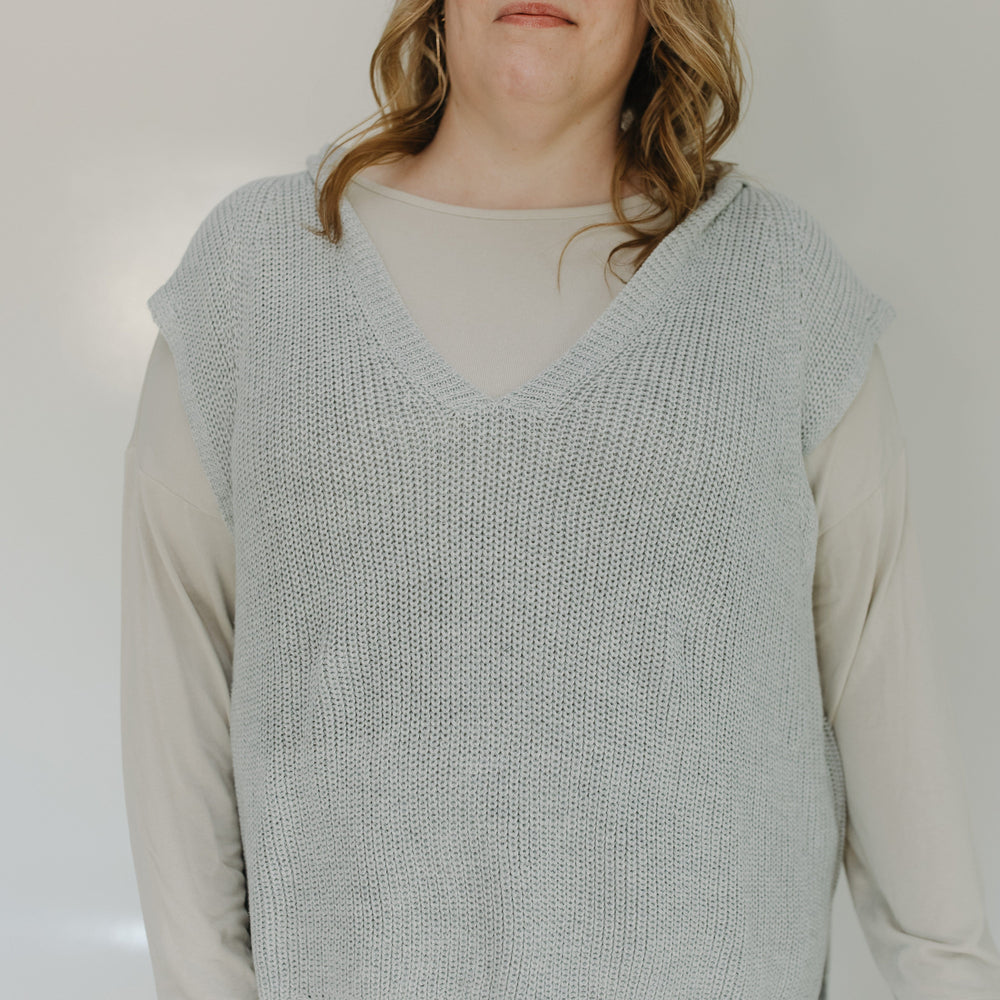 Person wearing a light gray knitted vest over a beige shirt with a plain background