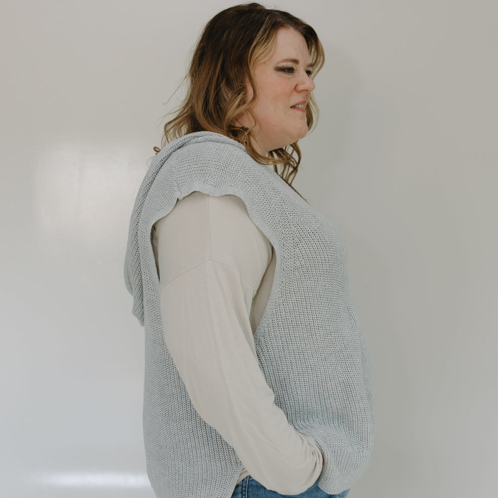 Person wearing a light blue knitted vest over a white shirt with blue jeans against a plain background
