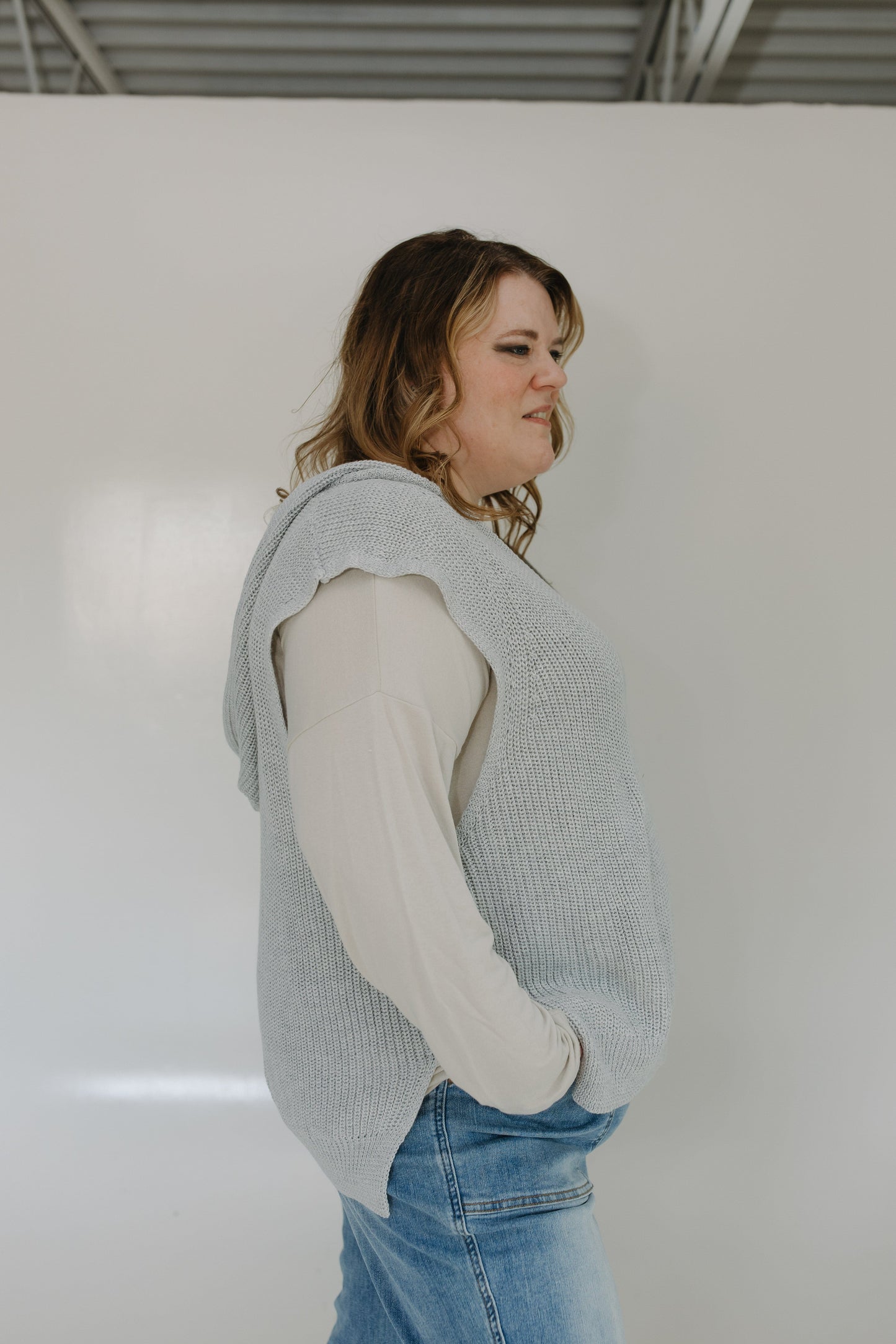 Person wearing a light blue knitted vest over a white shirt with blue jeans against a plain background