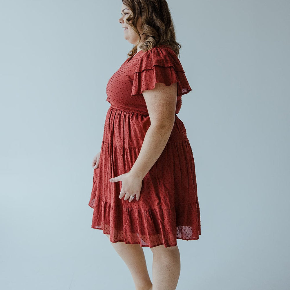 A woman in a Dex "Swiss Dot Double Flutter Sleeve Dress" in Spiced Cranberry with a tiered skirt stands in profile facing left, with wavy hair and wearing sandals, against a plain light-colored background.