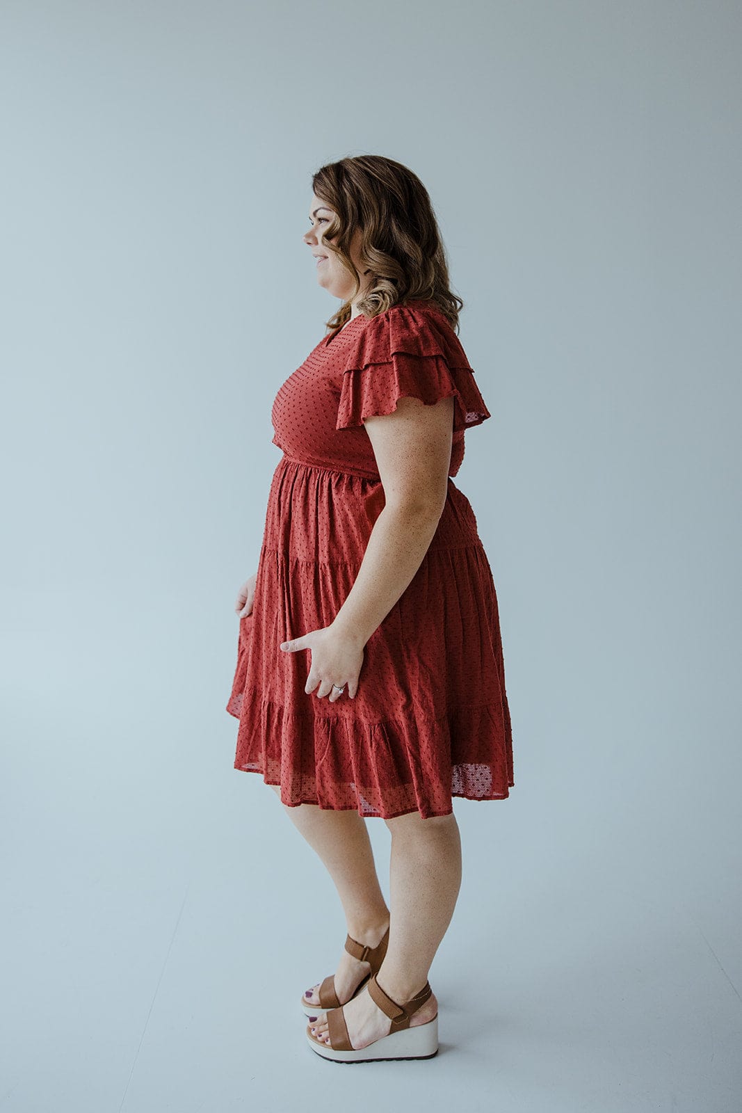 A woman in a Dex "Swiss Dot Double Flutter Sleeve Dress" in Spiced Cranberry with a tiered skirt stands in profile facing left, with wavy hair and wearing sandals, against a plain light-colored background.