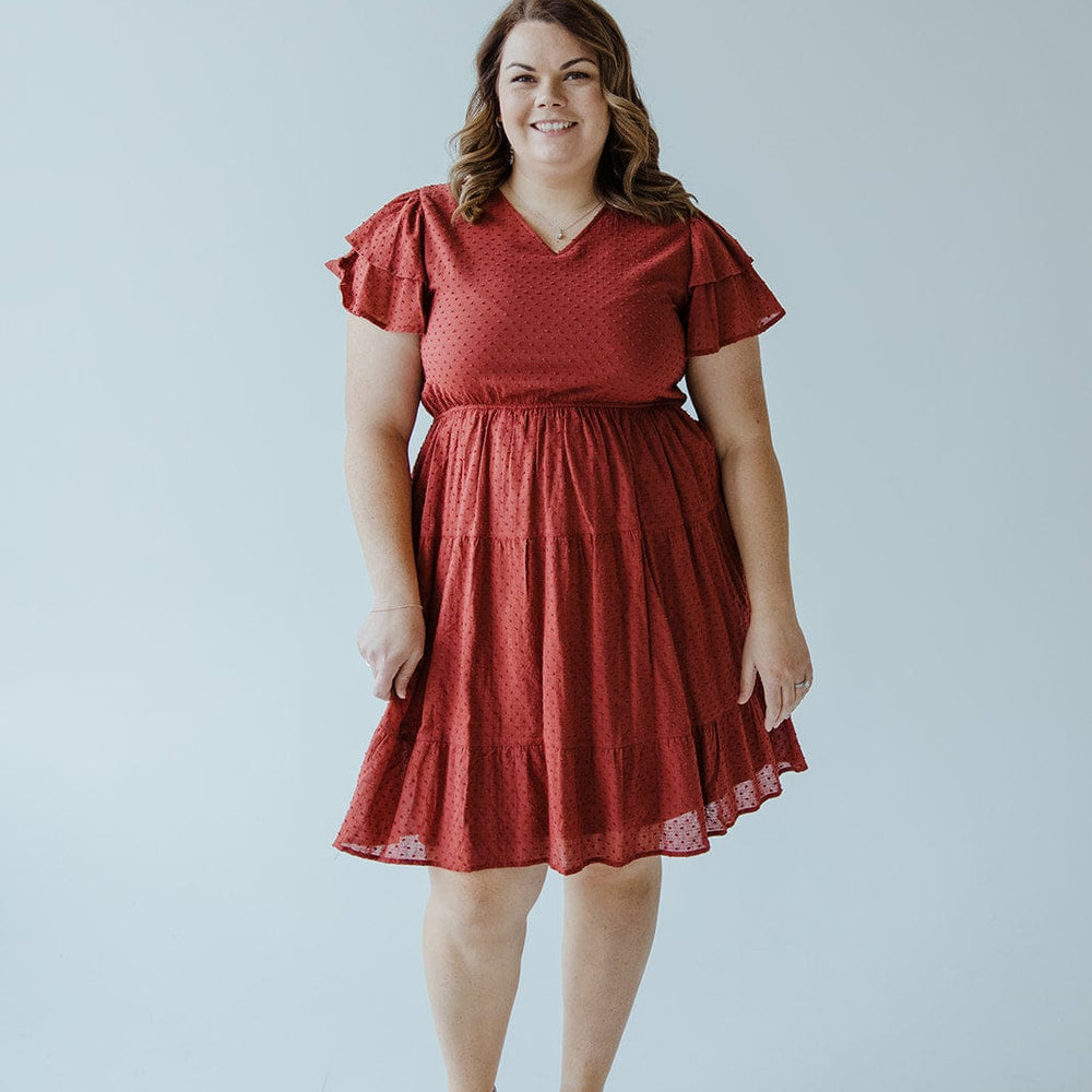 A person stands smiling, wearing Dex's SWISS DOT DOUBLE FLUTTER SLEEVE DRESS IN SPICED CRANBERRY with tan wedge sandals, against a plain light background.