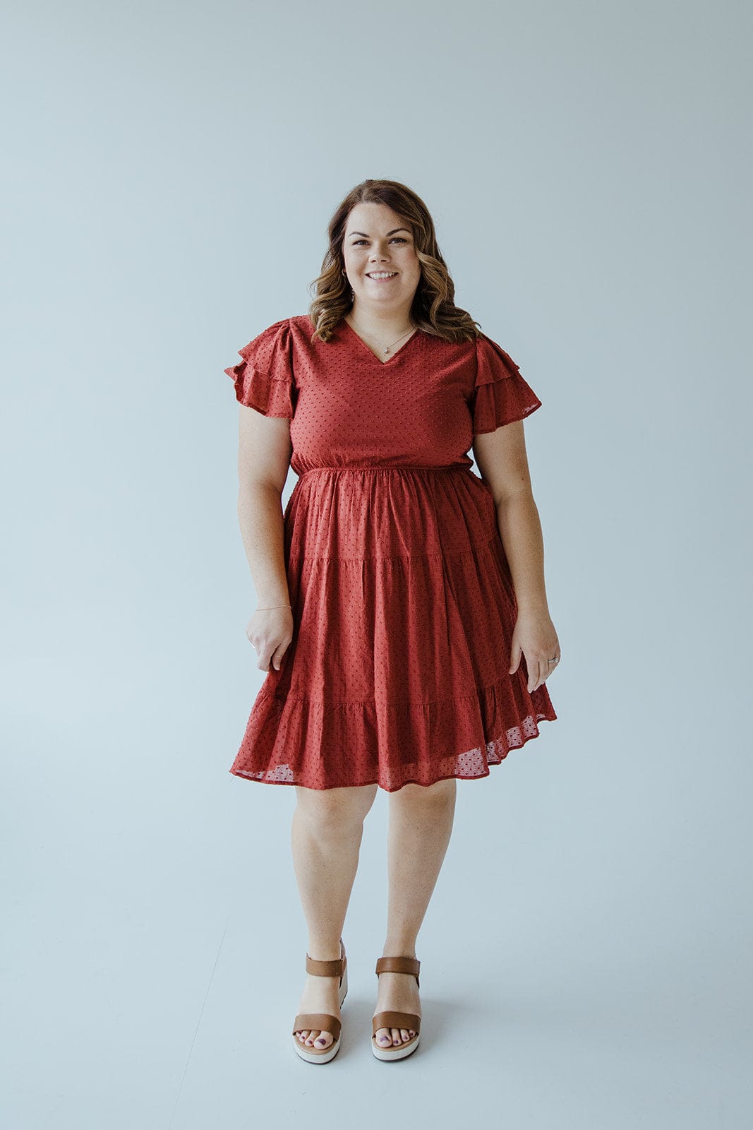 A person stands smiling, wearing Dex's SWISS DOT DOUBLE FLUTTER SLEEVE DRESS IN SPICED CRANBERRY with tan wedge sandals, against a plain light background.