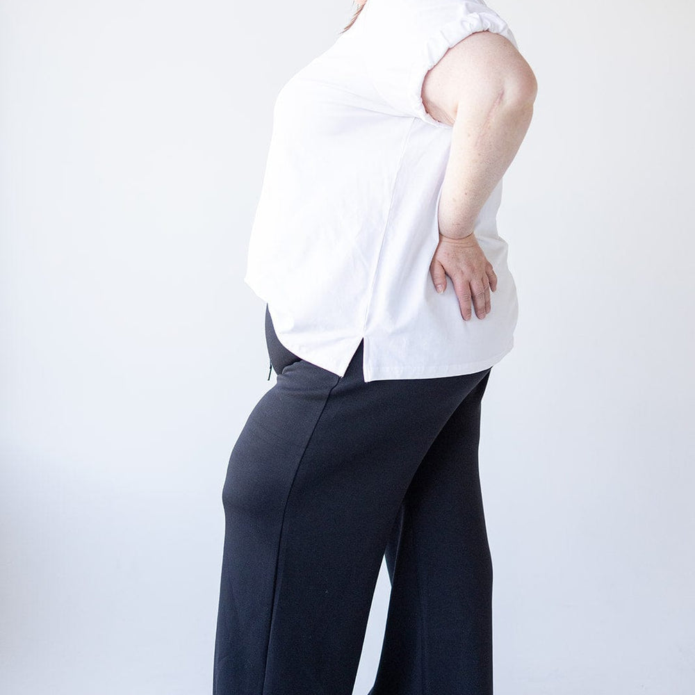 A person in a white shirt and black pants, featuring a relaxed fit, stands in profile against a plain background.