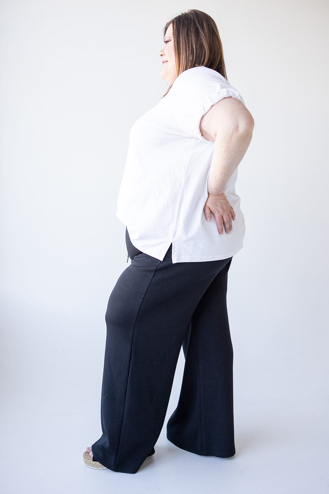 A person in a white shirt and black pants, featuring a relaxed fit, stands in profile against a plain background.