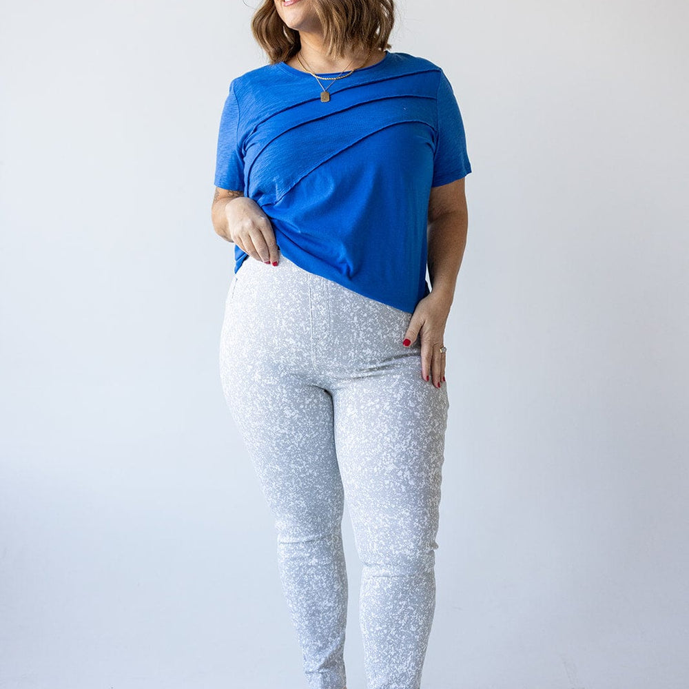 A woman in a top from Love Marlow and Spanx® Ankle Jean-ish® Leggings in Speckled Grey stands smiling against a plain backdrop. Her hand is in her pocket, and she is wearing beige flats with sunglasses perched on her head.