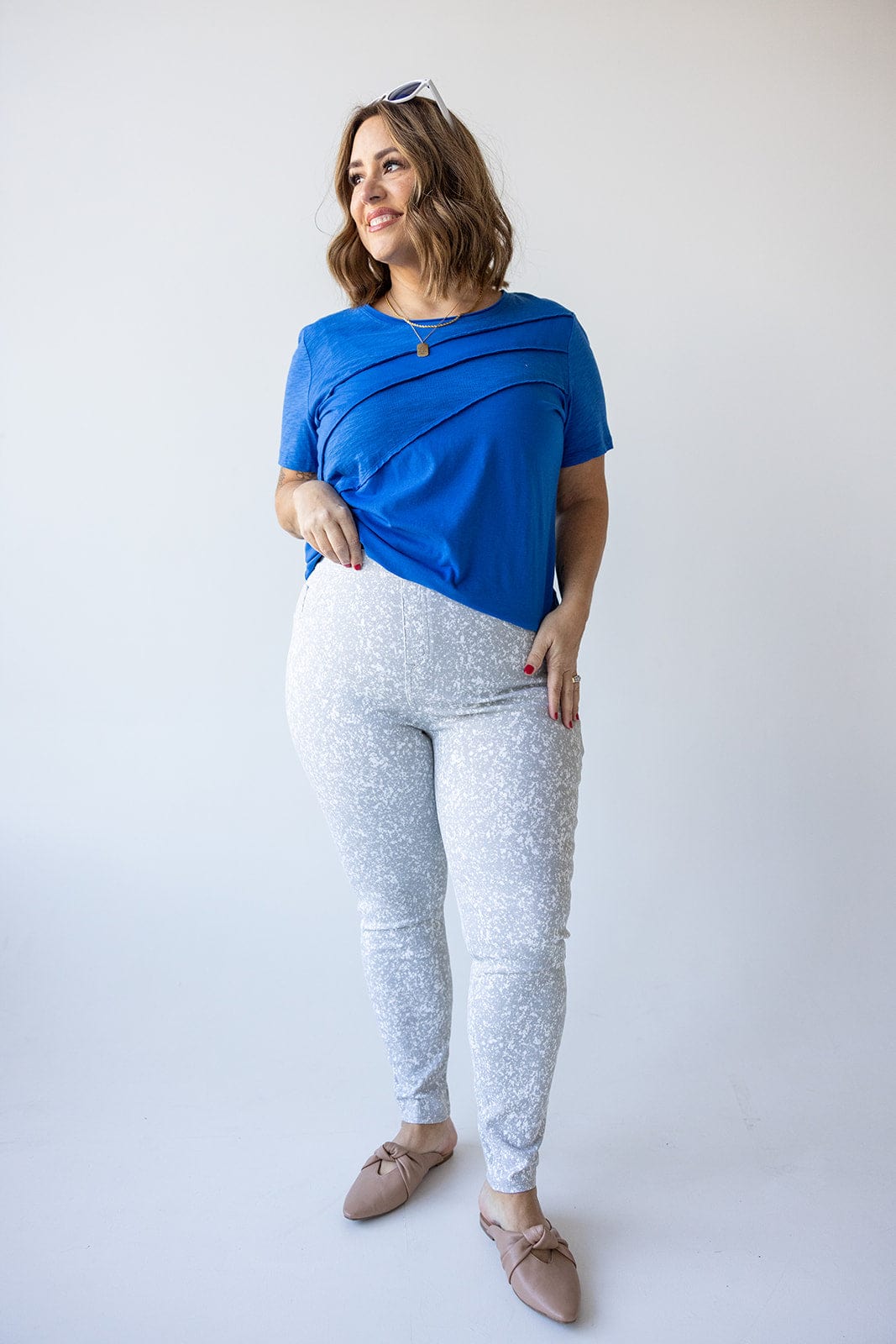 A woman in a top from Love Marlow and Spanx® Ankle Jean-ish® Leggings in Speckled Grey stands smiling against a plain backdrop. Her hand is in her pocket, and she is wearing beige flats with sunglasses perched on her head.