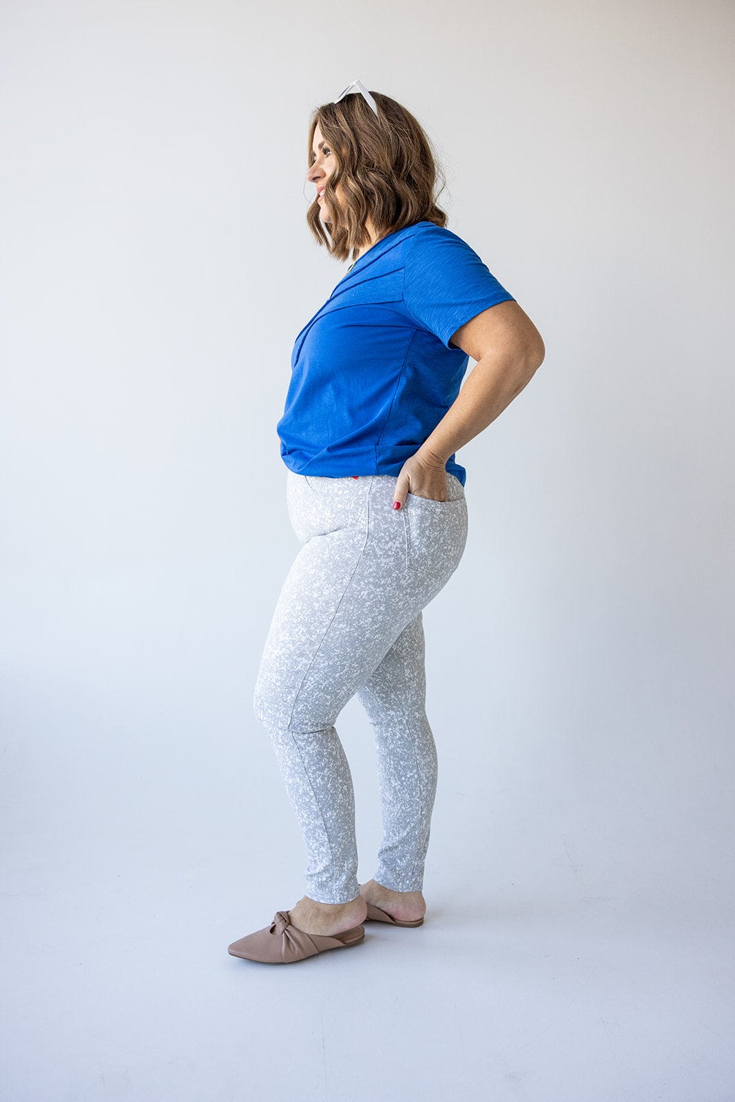 A person wearing a blue shirt and Spanx© Ankle Jean-ish® Legging in Speckled Grey, featuring tummy control, stands sideways against a white background.