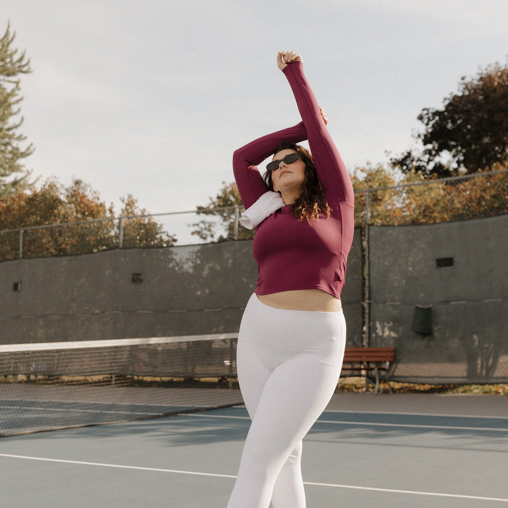 Person in maroon long-sleeve top and white leggings stretching on a tennis court. SPANX at Love Marlow Sioux Falls