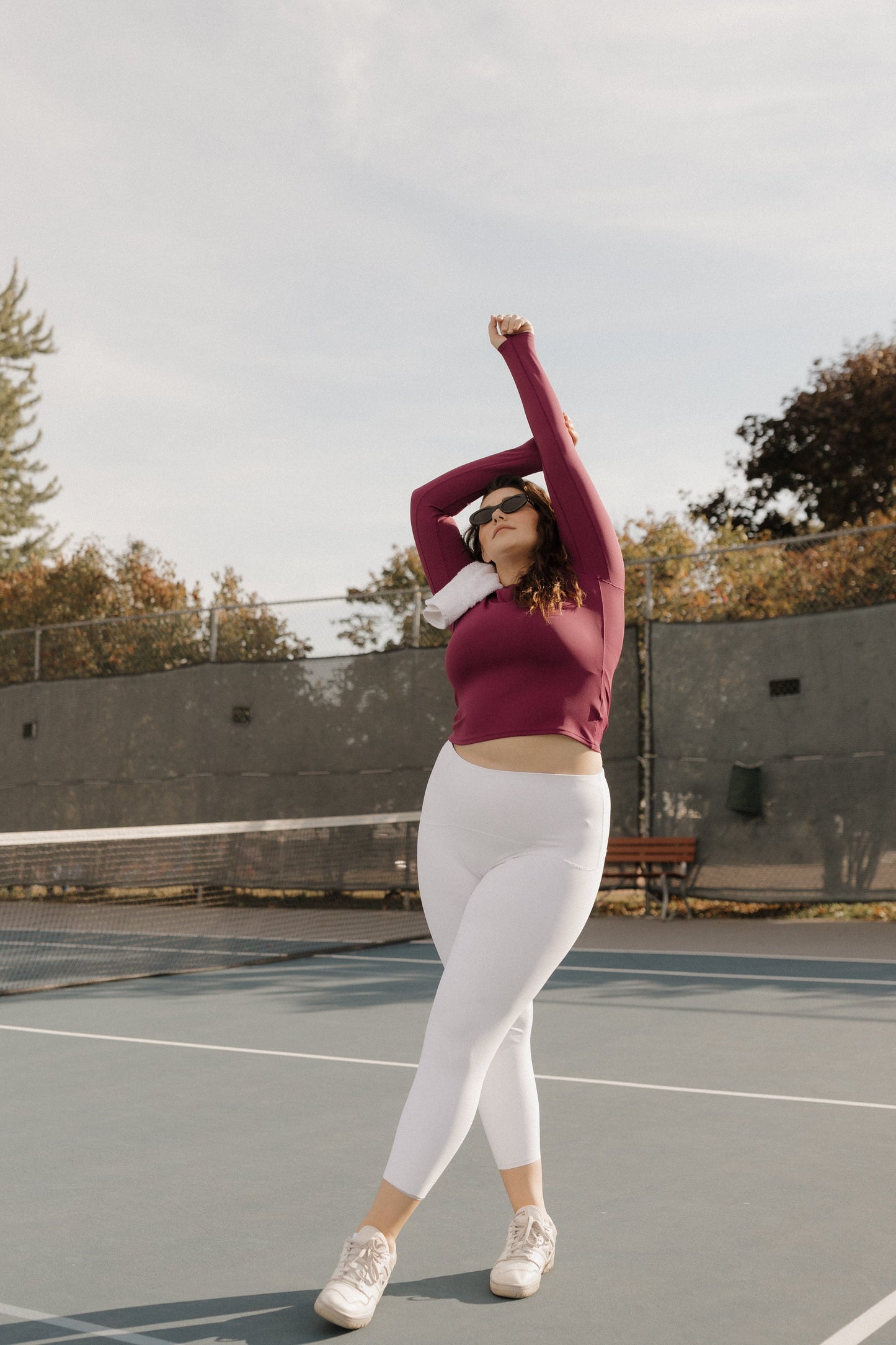 Person in maroon long-sleeve top and white leggings stretching on a tennis court. SPANX at Love Marlow Sioux Falls