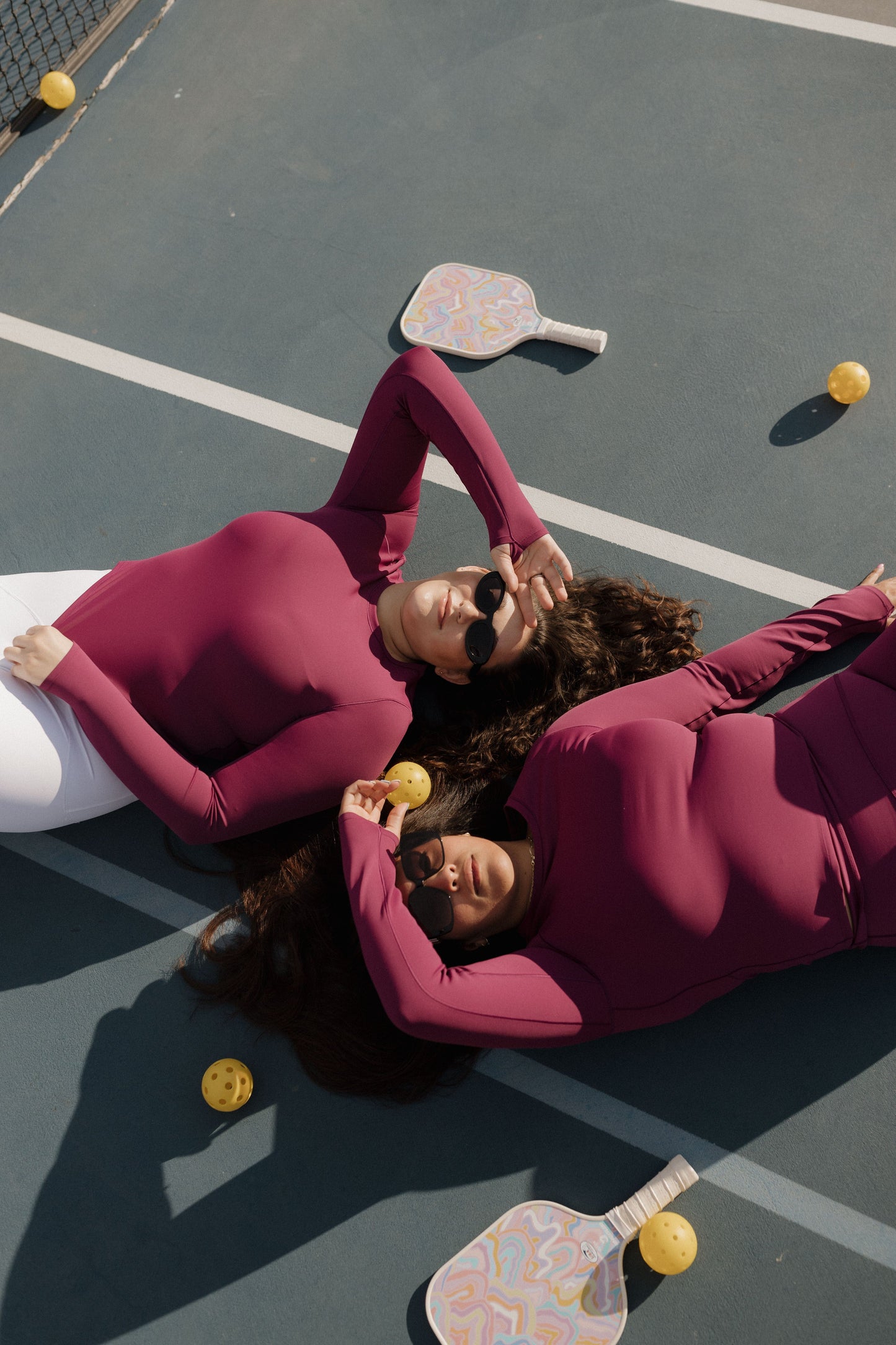 Two people lying on a tennis court with pickleball equipment and balls. SPANX activewear at Love Marlow in Sioux Falls