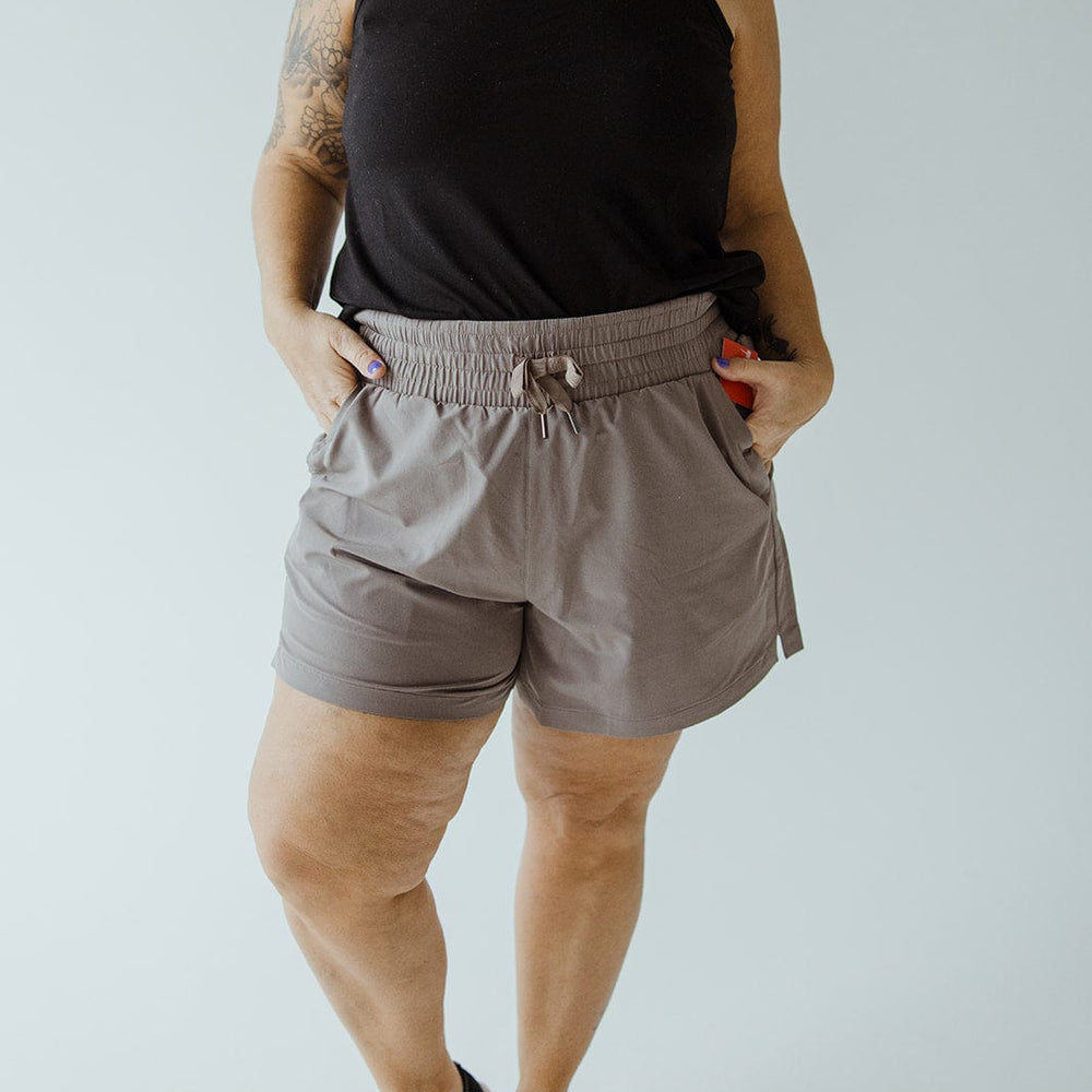 A woman wearing a black tank top, Spanx® CASUAL FRIDAYS SHORT IN SMOKE that offer high-rise coverage, and black sneakers stands with her hands in her pockets against a plain background.