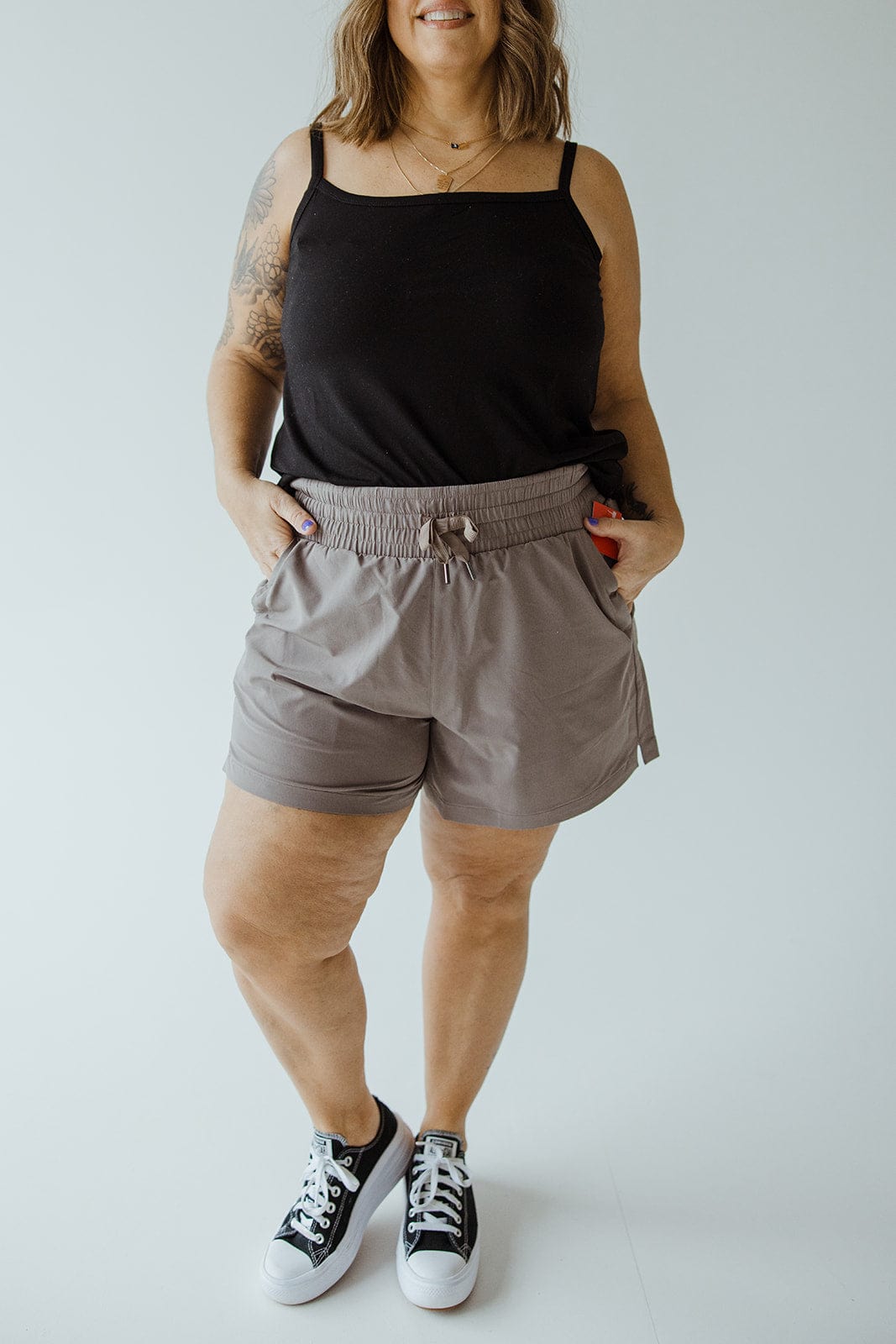 A woman wearing a black tank top, Spanx® CASUAL FRIDAYS SHORT IN SMOKE that offer high-rise coverage, and black sneakers stands with her hands in her pockets against a plain background.