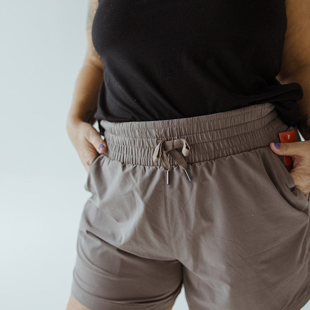 Person wearing a black sleeveless top and Spanx® CASUAL FRIDAYS SHORT IN SMOKE with high-rise coverage, their hands in the pockets, standing in front of a plain background.