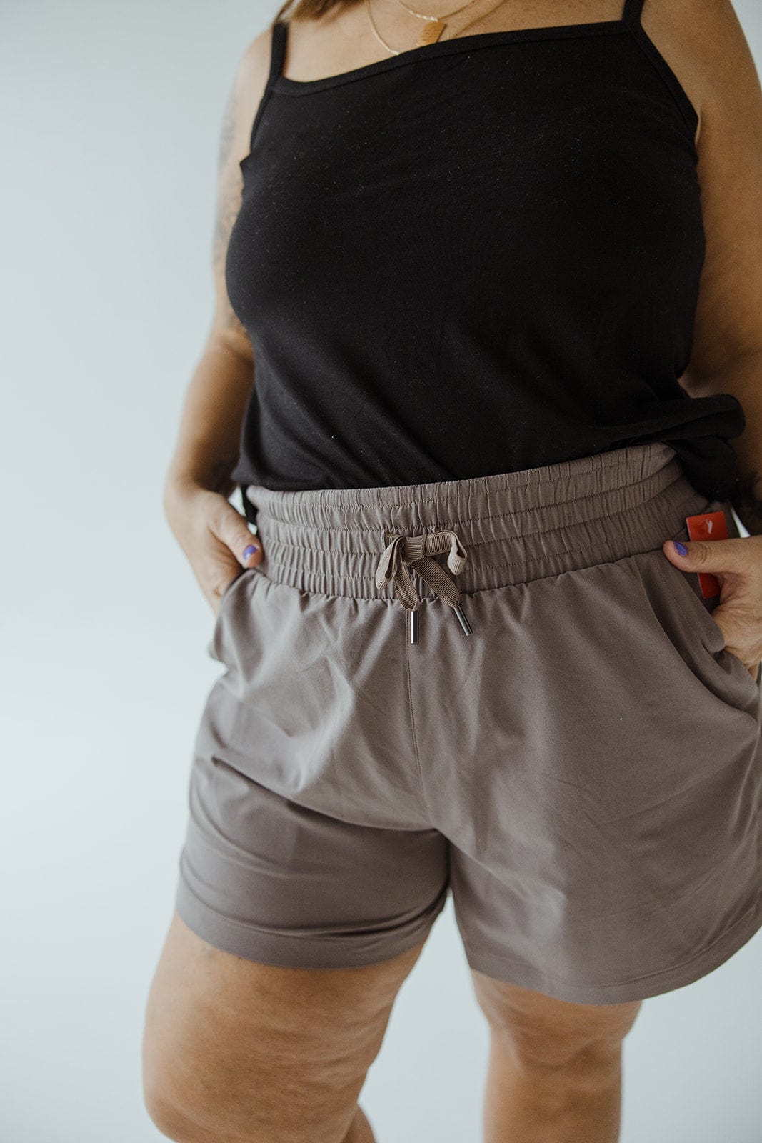 Person wearing a black sleeveless top and Spanx® CASUAL FRIDAYS SHORT IN SMOKE with high-rise coverage, their hands in the pockets, standing in front of a plain background.