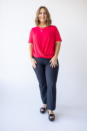 In front of a white backdrop, an individual is dressed in a red top paired with Spanx® Sunshine Kick Flare Pant in Very Black, which features 4-way stretch for enhanced comfort. Their look is complemented by black sandals, ideal for sunny days with UPF 50+ protection.