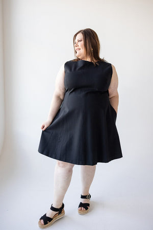 A woman stands against a plain white background, wearing the Spanx© THE PERFECT FIT AND FLARE DRESS IN BLACK crafted from smoothing premium ponte fabric and black sandals. Featuring a fit and flare shape, the dress also boasts functional pockets, as she looks to her left.