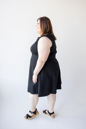 A woman in a sleeveless black dress made from smoothing premium ponte fabric stands in profile against a plain white background. She is wearing the Spanx© THE PERFECT FIT AND FLARE DRESS IN BLACK and black sandals.