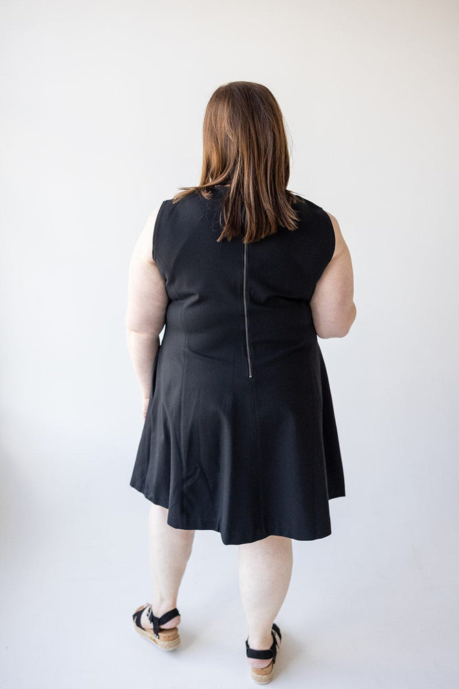 A person with long hair, wearing a sleeveless black dress in a fit and flare shape and sandals, is seen from the back against a plain white background. They are wearing the Spanx© THE PERFECT FIT AND FLARE DRESS IN BLACK by Spanx.