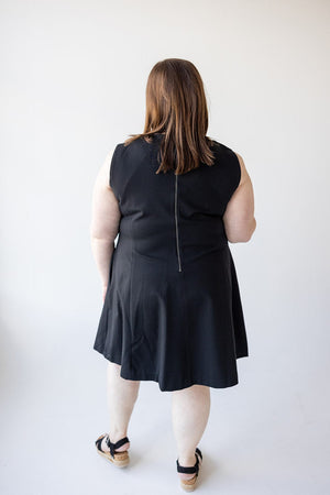 A person with long hair, wearing a sleeveless black dress in a fit and flare shape and sandals, is seen from the back against a plain white background. They are wearing the Spanx© THE PERFECT FIT AND FLARE DRESS IN BLACK by Spanx.