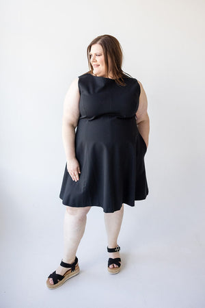 A person stands against a plain white background wearing Spanx© THE PERFECT FIT AND FLARE DRESS IN BLACK and stylish black sandals, looking slightly to the side.