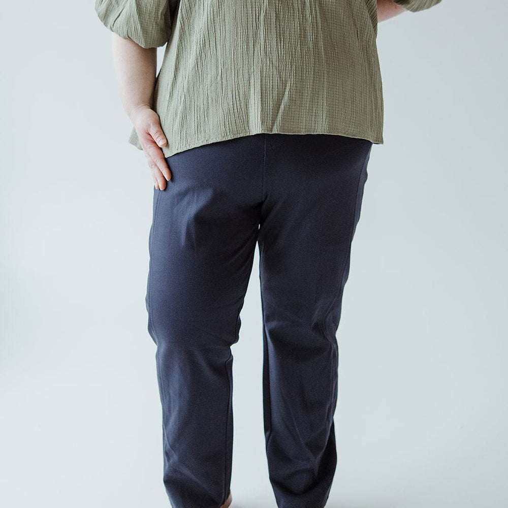 A person wearing a green top, Spanx© The Perfect Black Pant, Slim Straight in Classic Navy, and beige shoes stands against a plain white background.