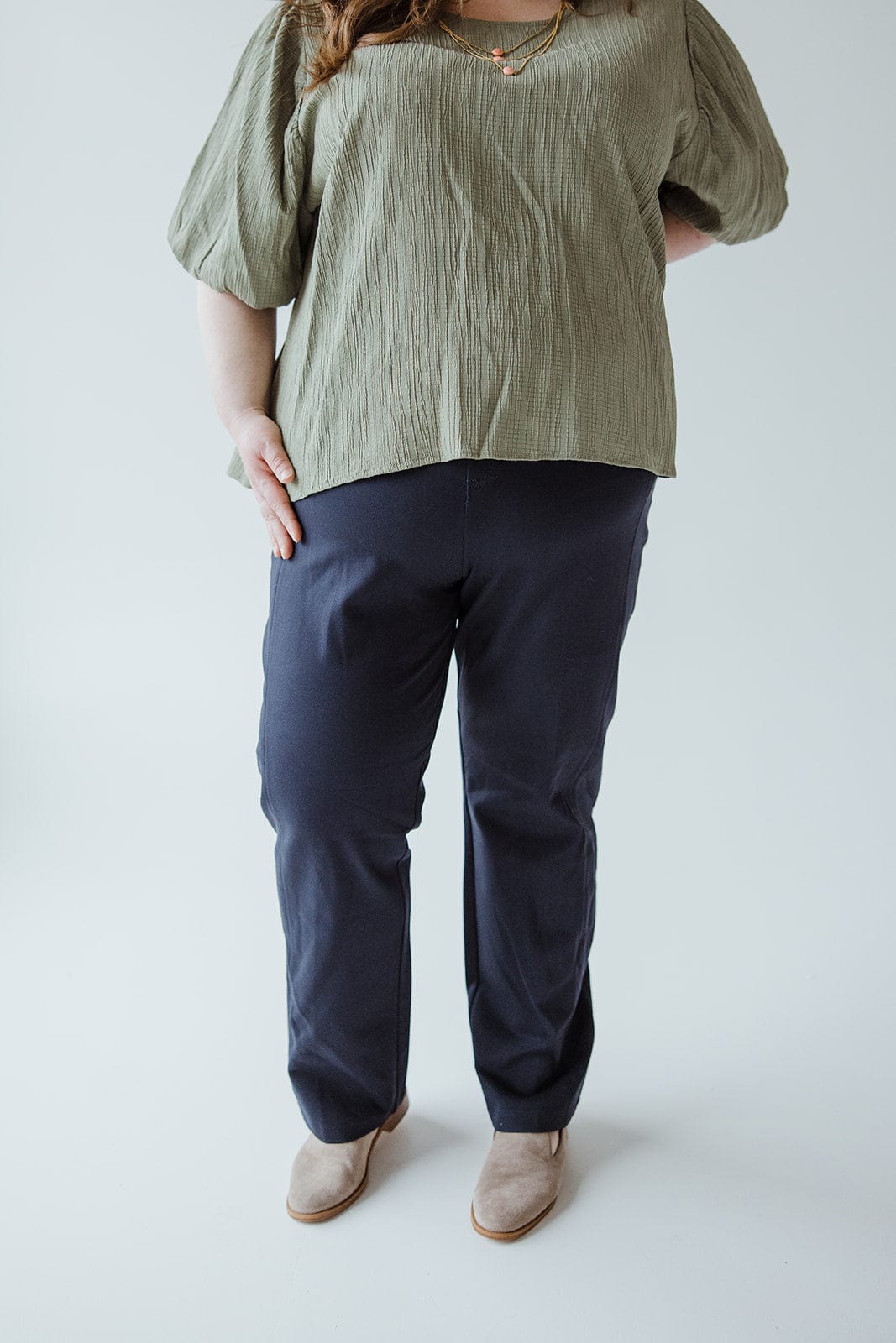 A person wearing a green top, Spanx© The Perfect Black Pant, Slim Straight in Classic Navy, and beige shoes stands against a plain white background.