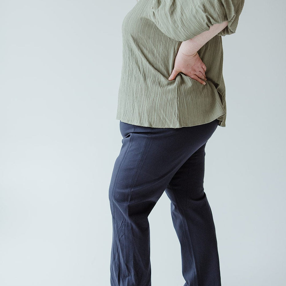 A person wearing a textured green top, Spanx© The Perfect Black Pant, Slim Straight in Classic Navy crafted from smoothing ponte fabric, and beige shoes stands against a plain background.