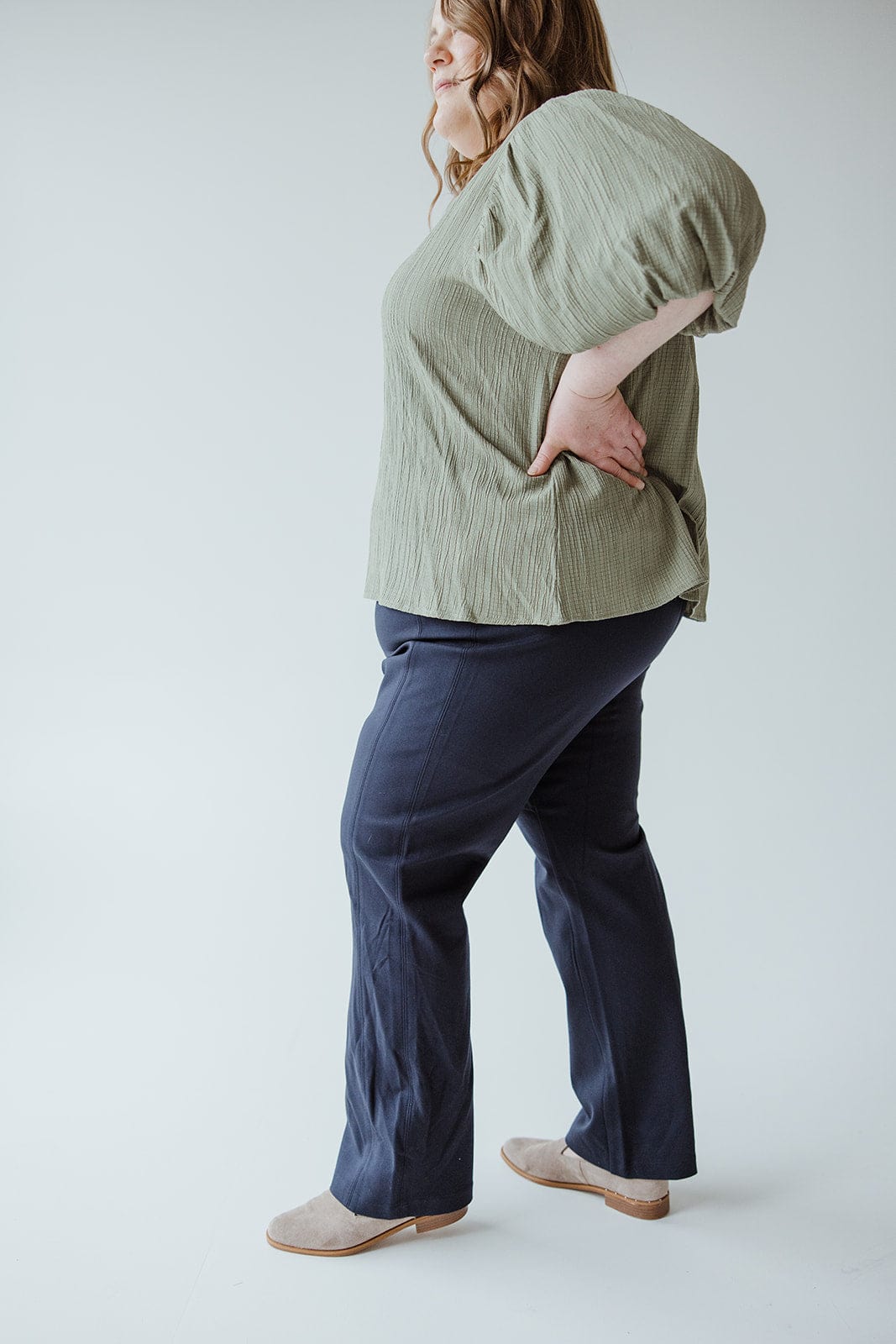 A person wearing a textured green top, Spanx© The Perfect Black Pant, Slim Straight in Classic Navy crafted from smoothing ponte fabric, and beige shoes stands against a plain background.