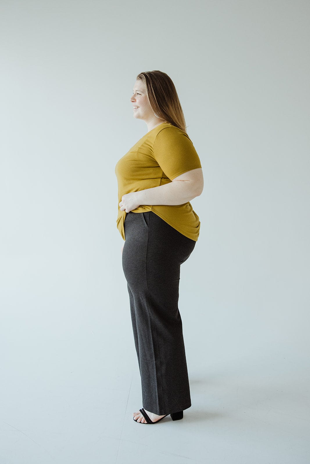 Person standing sideways, wearing a mustard yellow top and dark wide-leg pants made from premium ponte fabric, with a chic pull-on design. Black heels complete the look against a plain background.