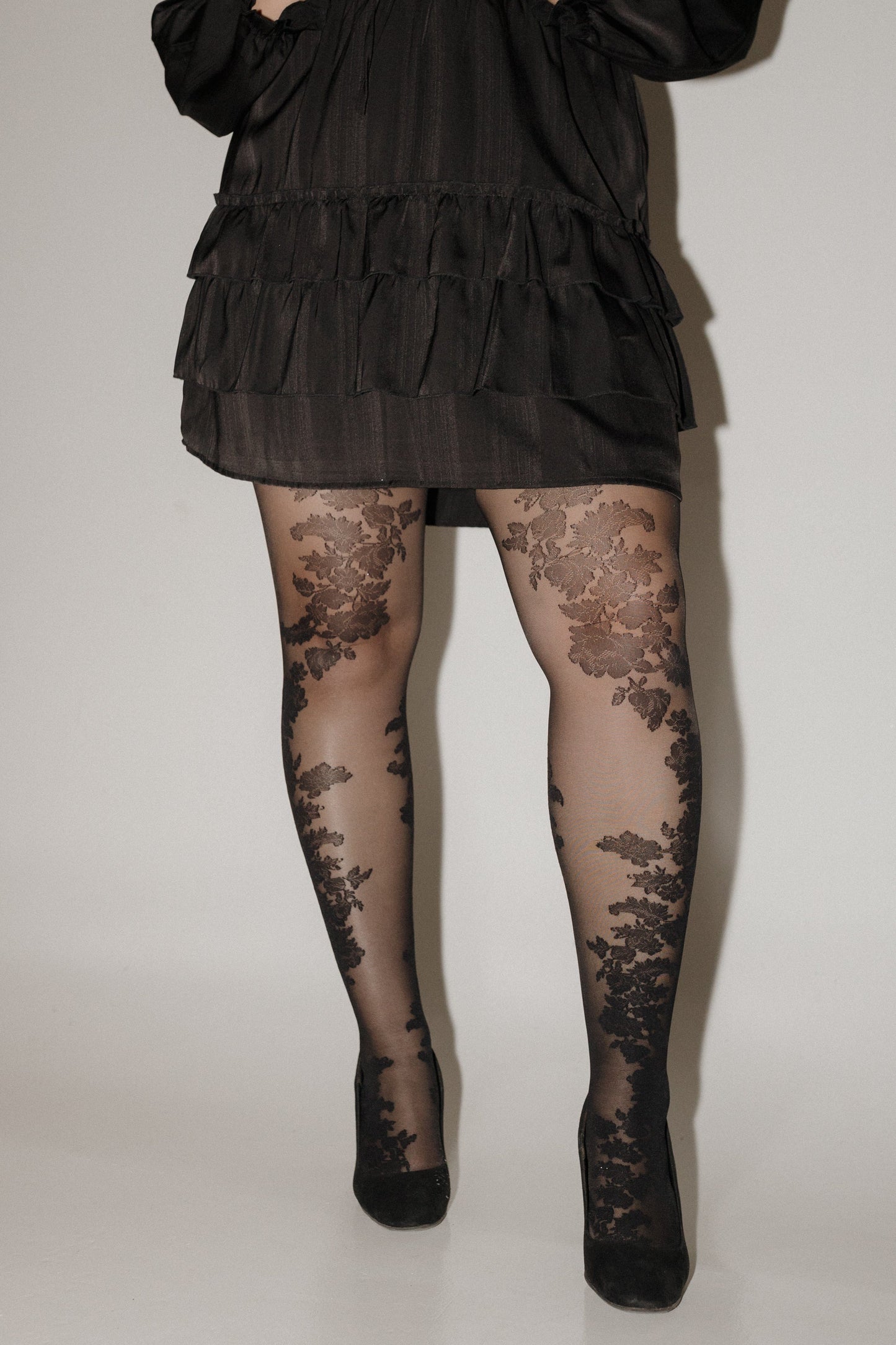 Person wearing a black ruffled dress and floral tights on a plain background SPANX floral tights