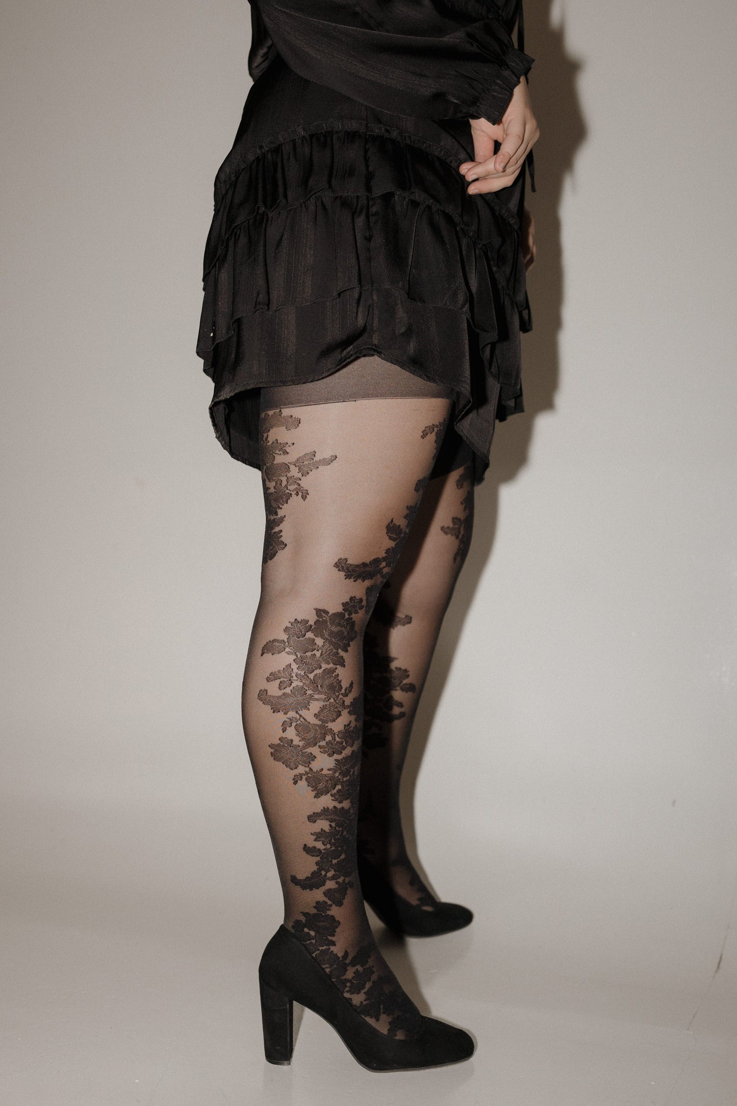 Person wearing a black dress with floral lace stockings and high heels on a plain background SPANX floral tights