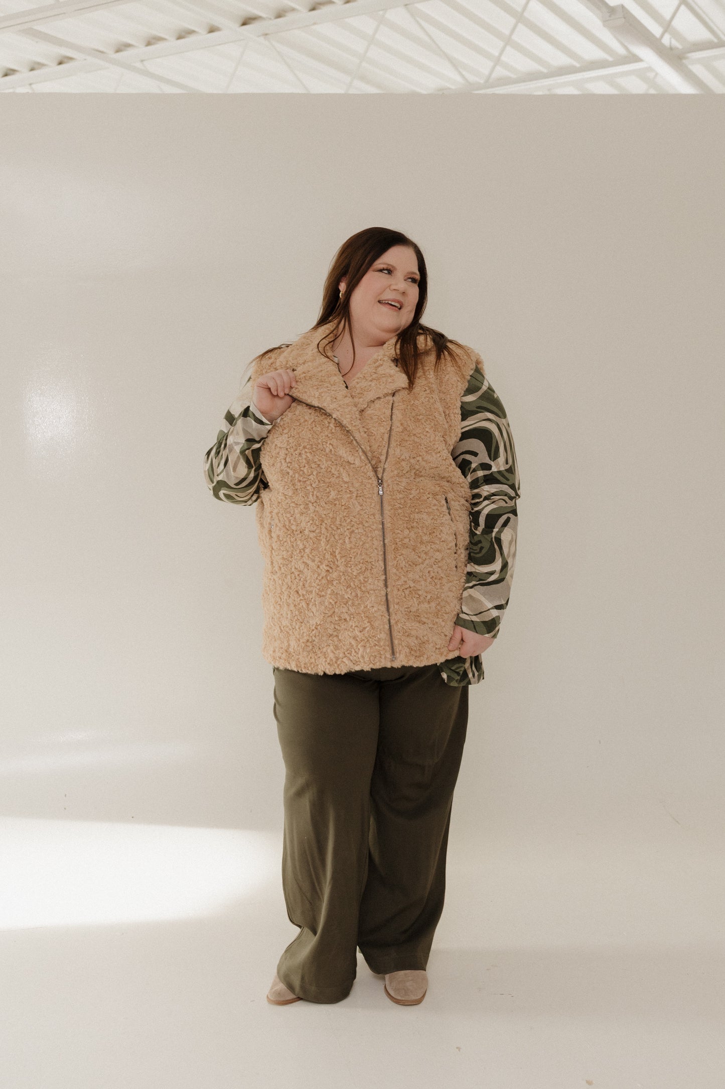 Person wearing a beige vest over a patterned shirt with green pants on a white background