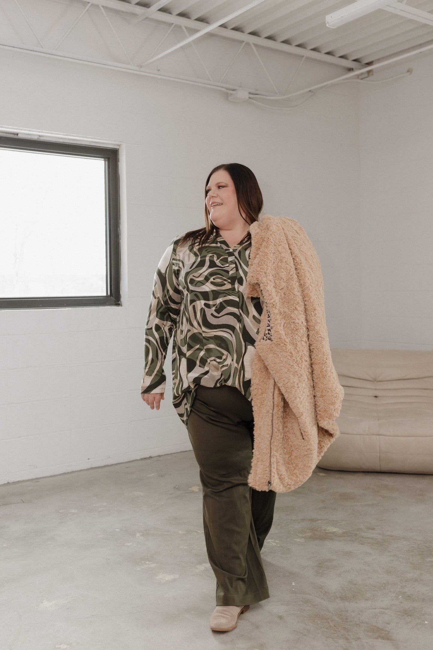 Person wearing a patterned shirt and beige coat in a minimalistic room.