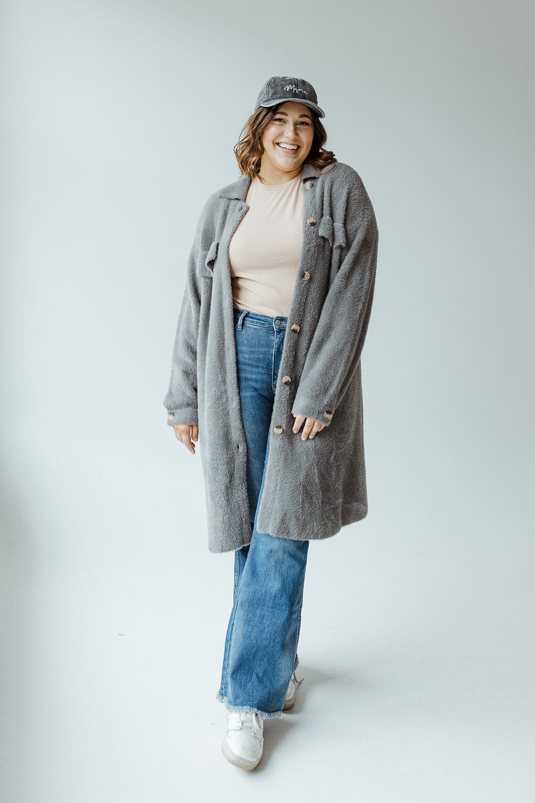 A person wearing a steel grey shacket, blue jeans, white sneakers, and a cap stands smiling against a plain backdrop.