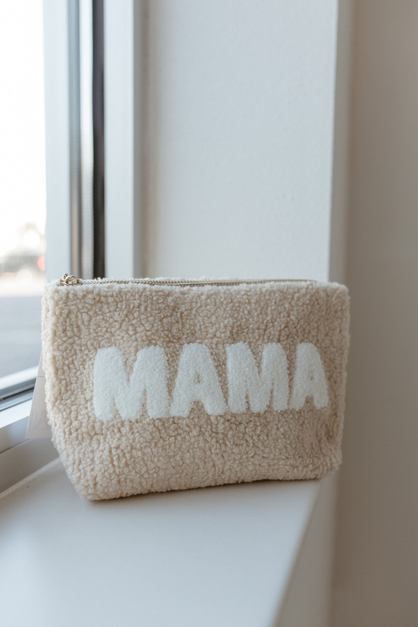 A beige pouch, named "MAMA Teddy Makeup Pouch," crafted from ultra-soft sherpa fabric with a retro floral interior lining, is placed on a windowsill.