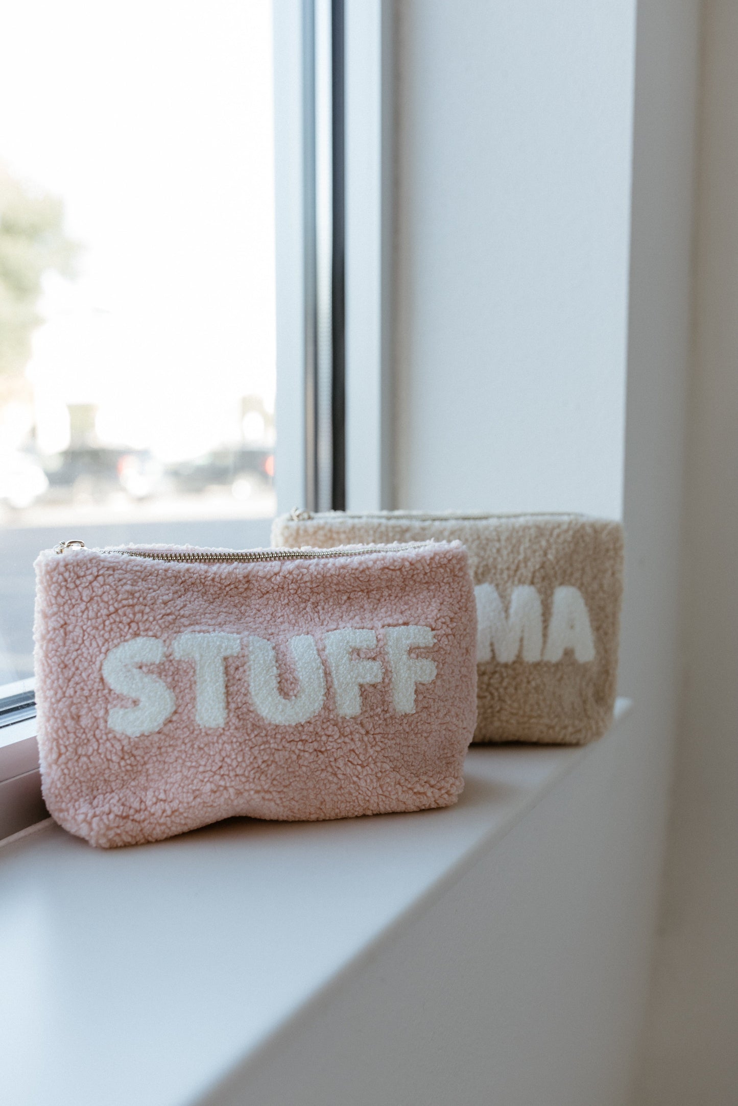 Two soft, textured pouches rest on a windowsill. The pink one reads "STUFF," while the beige one partially reveals "MA." Crafted from ultra-soft sherpa fabric, the MAMA Teddy Makeup Pouch boasts a retro floral interior lining, adding charm to its cozy exterior.