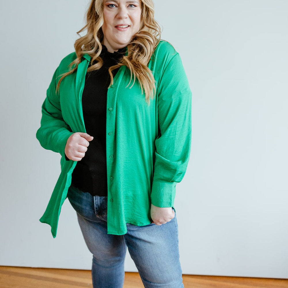 A woman with long, wavy hair wearing a TEXTURED BUTTON-UP BLOUSE IN EMERALD by Love Marlow, a black top, blue jeans, and black sneakers stands indoors on a wooden floor, with one hand in her pocket. Her professional appearance is complemented by the luxurious textured blouse she's sporting.