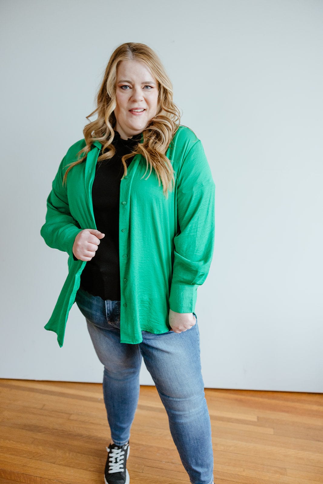 A woman with long, wavy hair wearing a TEXTURED BUTTON-UP BLOUSE IN EMERALD by Love Marlow, a black top, blue jeans, and black sneakers stands indoors on a wooden floor, with one hand in her pocket. Her professional appearance is complemented by the luxurious textured blouse she's sporting.