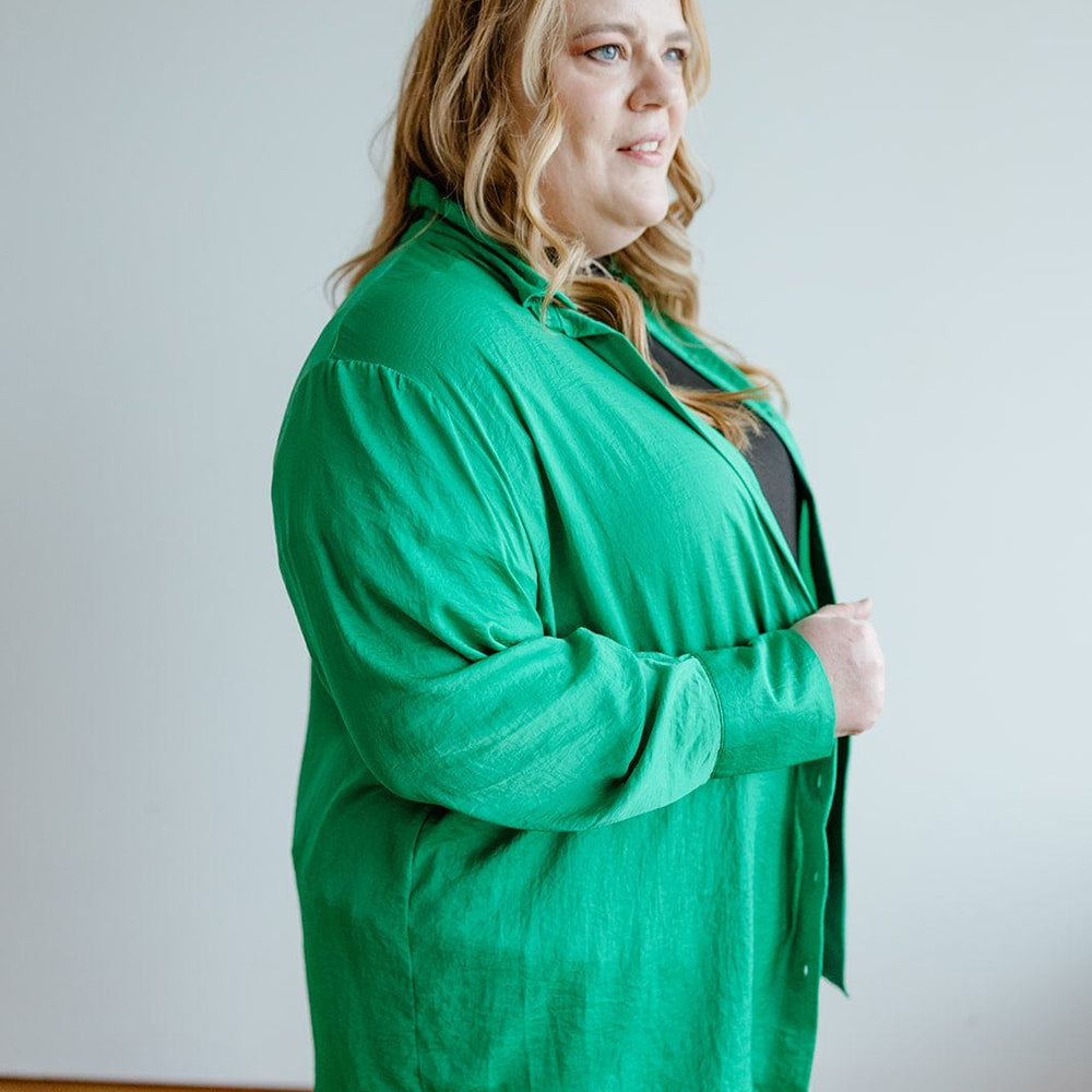 A woman with long, wavy blonde hair is wearing the luxurious TEXTURED BUTTON-UP BLOUSE IN EMERALD by Love Marlow, paired with blue jeans. She stands indoors against a plain, light-colored background, exuding a professional appearance.