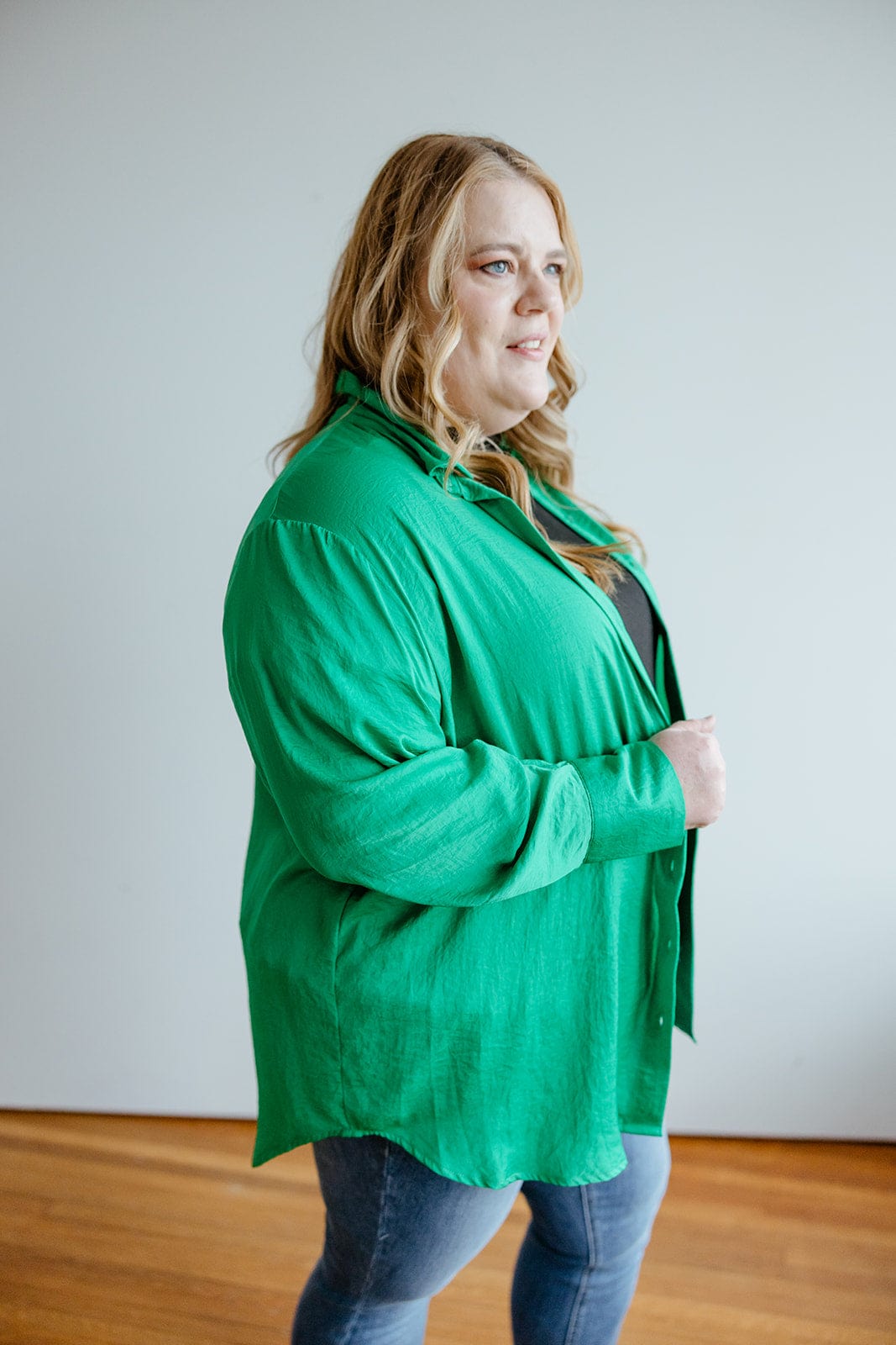 A woman with long, wavy blonde hair is wearing the luxurious TEXTURED BUTTON-UP BLOUSE IN EMERALD by Love Marlow, paired with blue jeans. She stands indoors against a plain, light-colored background, exuding a professional appearance.