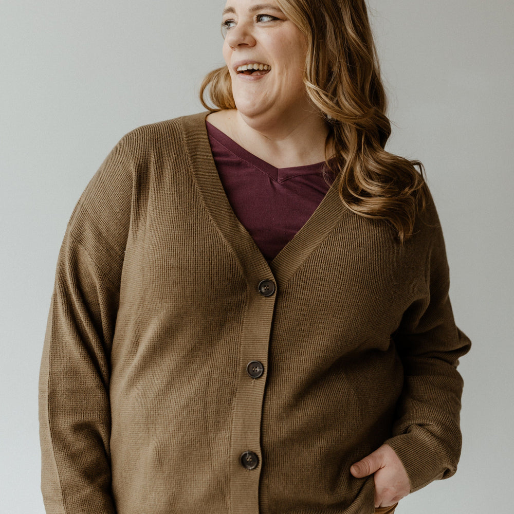 A person wearing a cozy, textured button-front cardigan stands with one hand in their pocket, looking to the side and smiling.