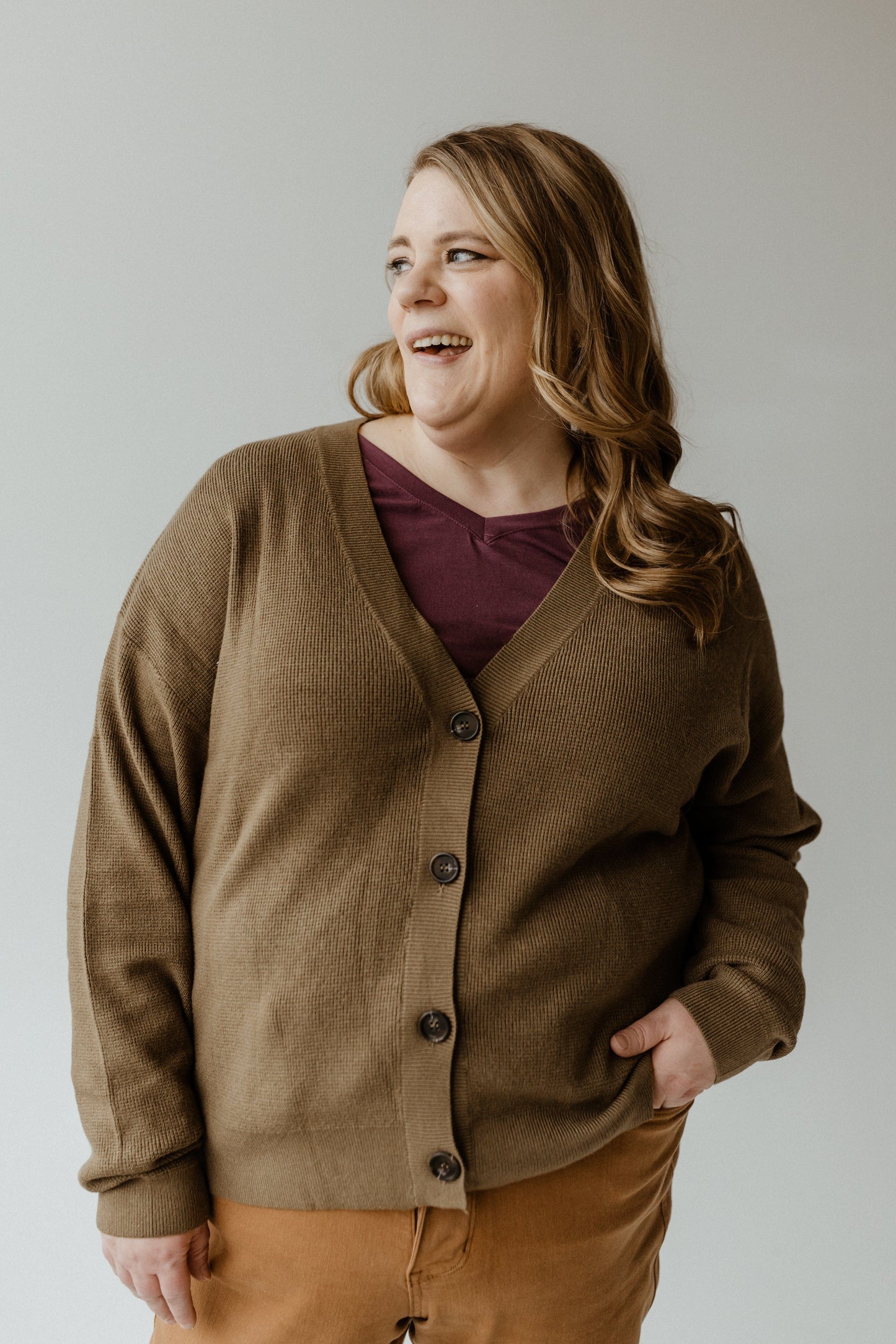 A person wearing a cozy, textured button-front cardigan stands with one hand in their pocket, looking to the side and smiling.