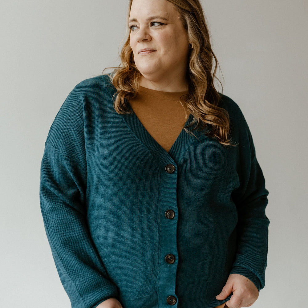 A person with long hair wearing a textured button-front cardigan in teal with ribbed hem and cuffs, paired with a mustard top, stands against a plain background, looking to the side.