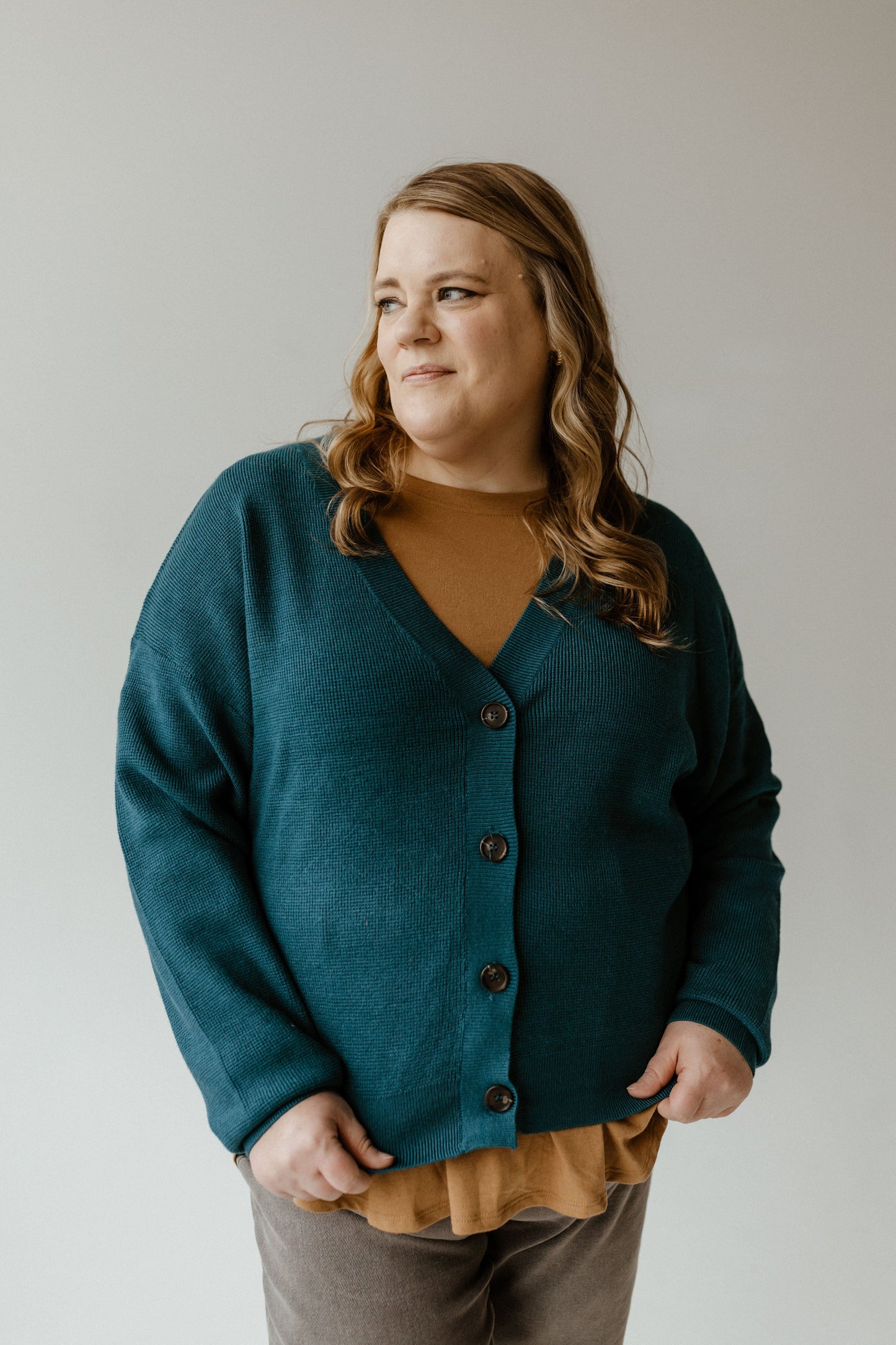 A person with long hair wearing a textured button-front cardigan in teal with ribbed hem and cuffs, paired with a mustard top, stands against a plain background, looking to the side.