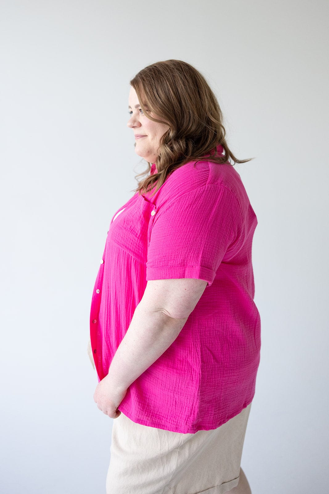 A person with long hair is standing against a plain white background, facing the left side, wearing a bright pink Yesta TEXTURED COTTON BUTTON UP BLOUSE IN AZALEA.