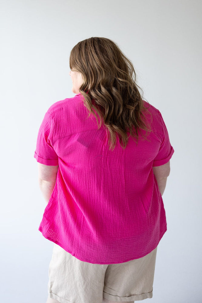 A person with long hair stands with their back to the camera, wearing a bright pink "Textured Cotton Button Up Blouse in Azalea" by Yesta and beige shorts made from textured cotton fabric against a plain background.